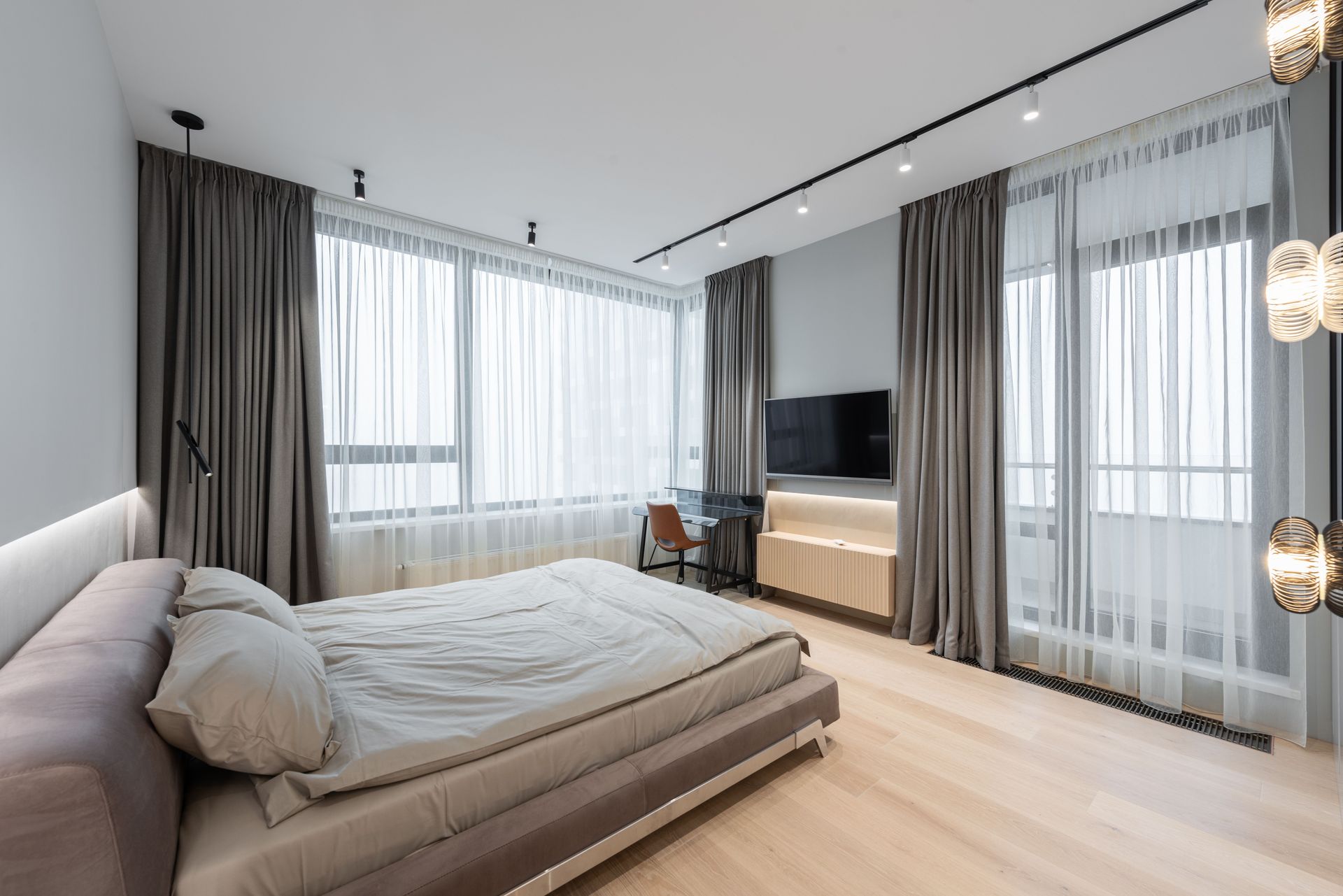 Interior of modern light bedroom with bed next to TV with curtains on windows and table with chair